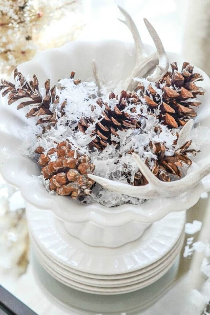 DECORATING WITH RISERS-stack of white dishes with a bowl filled with pinecones and faux snow