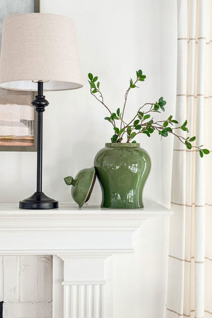 Winter mantel decorated with a green ceramic jar, simple greenery, and a table lamp beside artwork