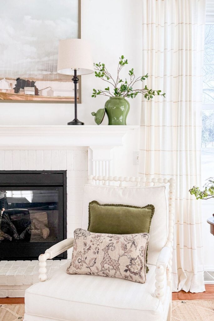 Winter mantel decorated with greenery in a ceramic jar, a table lamp, and simple neutral accents beside a fireplace