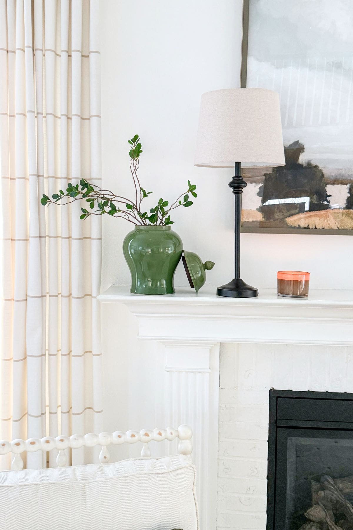 Winter mantel side decorated with a table lamp, greenery in a ceramic jar, and simple neutral accents