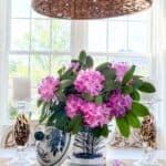 Blue and white chinoiserie planter filled with pink rhododendron blooms on a dining table beneath a woven pendant light, styled with glass candlesticks in a bright summer dining room.