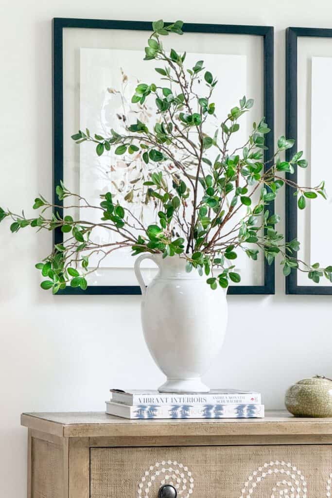 White ceramic vase filled with greenery branches styled on books atop a dresser for budget friendly home decorating.
