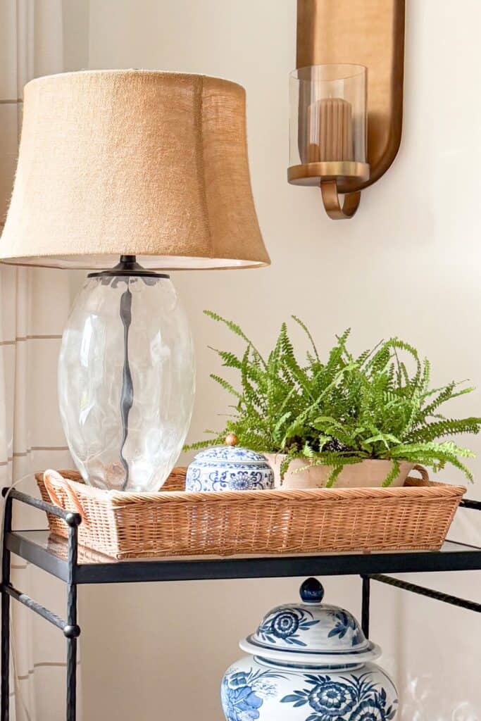 Decorative basket tray styled with a glass lamp, fern, and blue and white ginger jar on a black accent table for budget friendly home decorating.