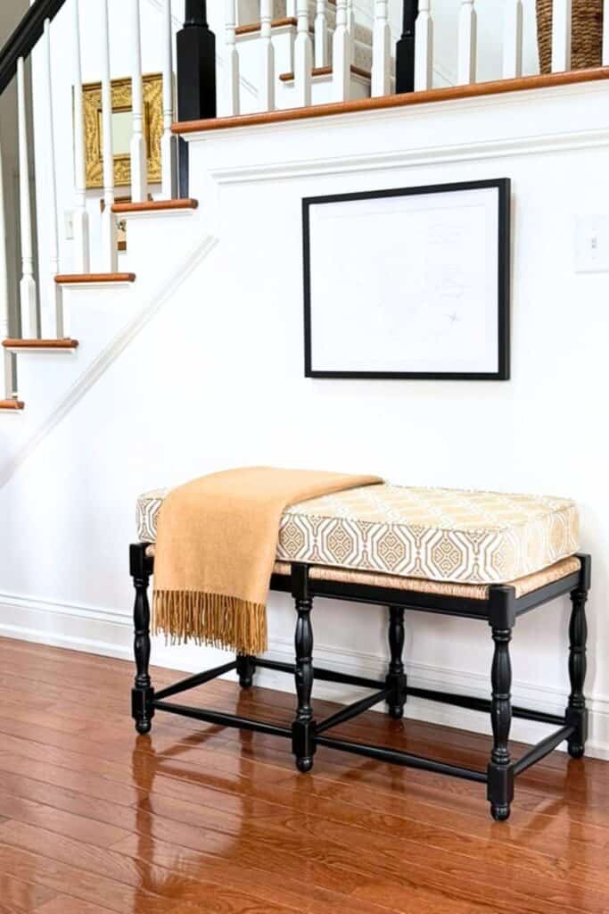 Black bench with a patterned cushion and camel throw beneath framed artwork in a neutral foyer, showing how black accents add visual weight.