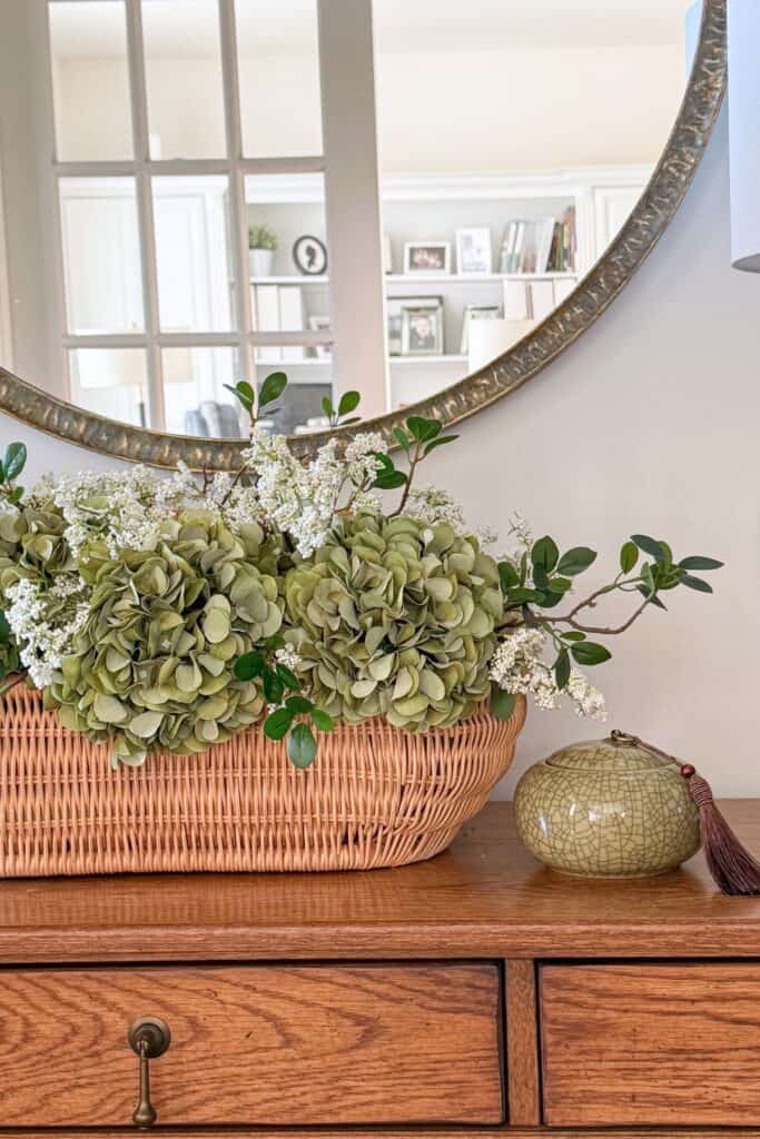 Wicker basket filled with green hydrangeas and greenery styled on a wooden console table beneath a round mirror beside a blue and white lamp.