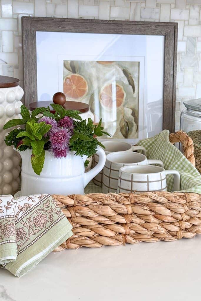 Woven basket styled with mugs, flowers, a tea towel, and kitchen accessories on a countertop for budget friendly decorating.