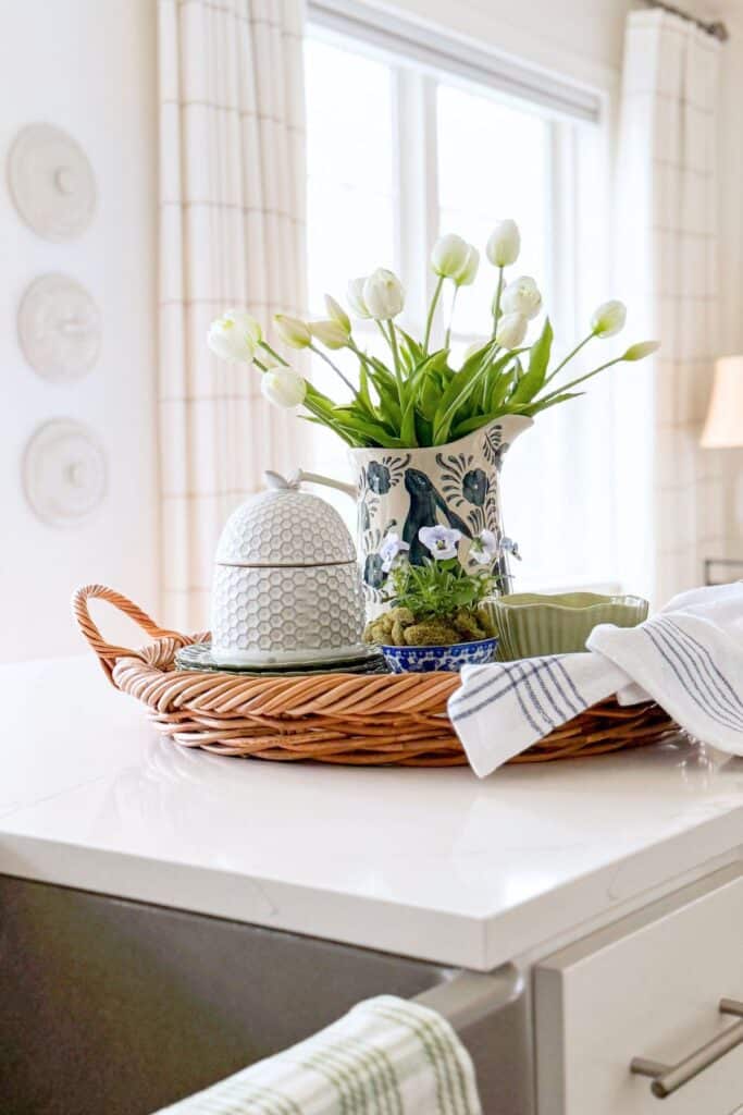 Wicker tray styled with white tulips, dishes, and a tea towel on a kitchen island for budget friendly decorating.