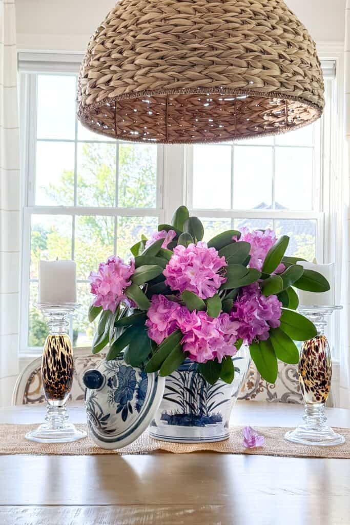 Pink rhododendron blooms arranged in a blue and white planter on a dining room table beneath a woven pendant light.