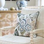 Cream upholstered chair with floral pillow styled beside a blue and white lamp and wood chest in a budget friendly transitional living room.