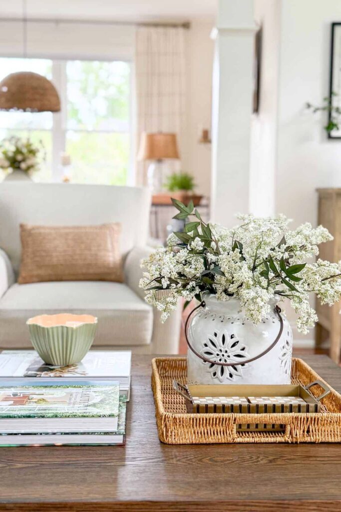 White ceramic vase with white flowers and green stems styled on a wicker tray on a coffee table with stacked books and a small green bowl