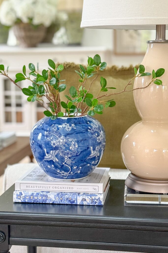 Blue and white chinoiserie vase with green stems styled on stacked books on a dark end table beside a neutral ceramic lamp