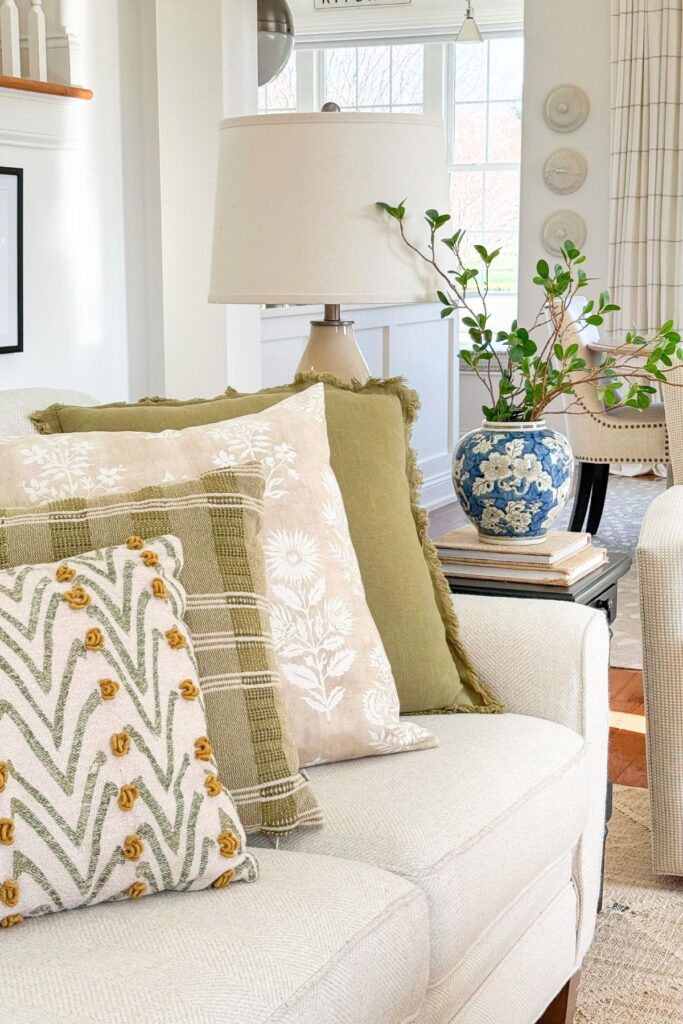 Green and neutral patterned throw pillows layered on a white sofa in a bright living room with a blue and white ginger jar vase