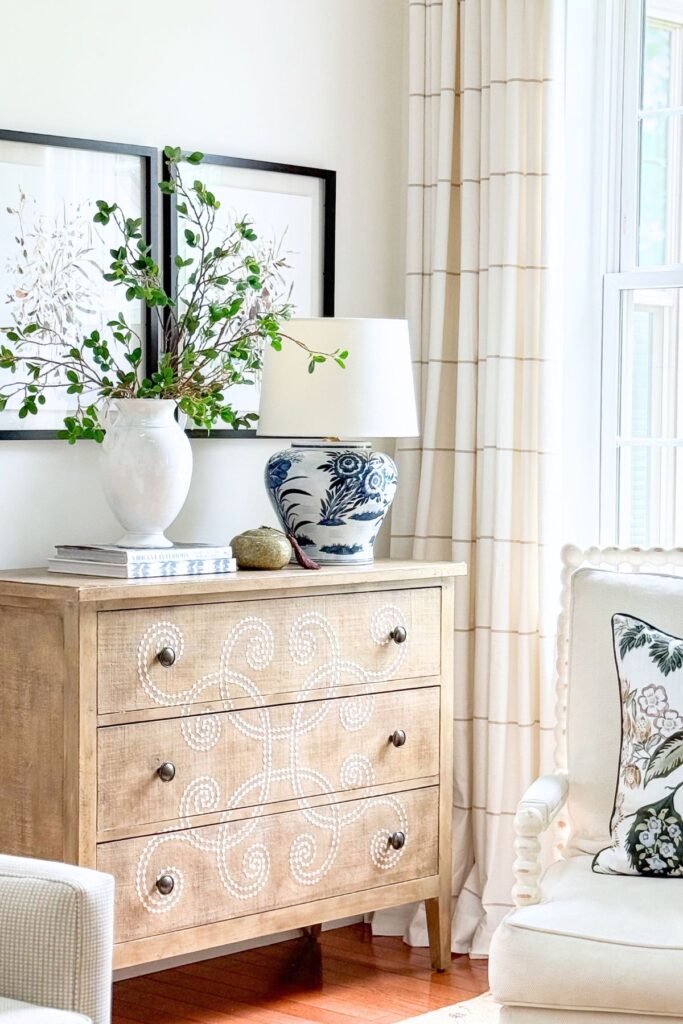 Neutral wood chest with carved detail styled with a white vase of green branches, a blue and white ginger jar lamp, and stacked books beside framed botanical art