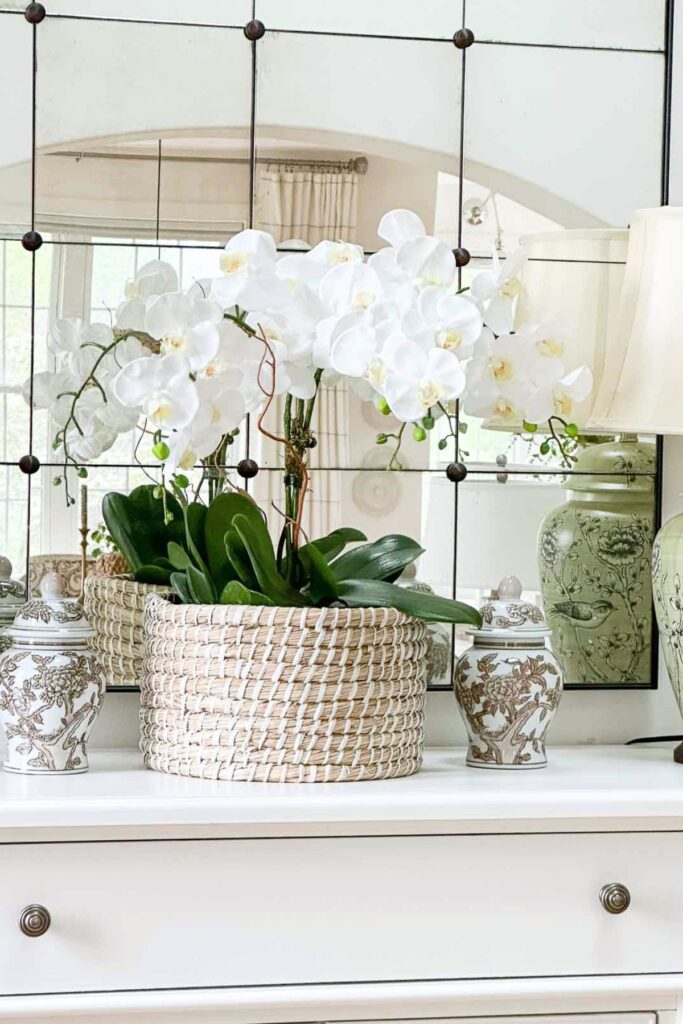 White orchid plant in a woven basket styled on a white dresser with small ginger jar accents and a green ceramic lamp beside an antique mirror