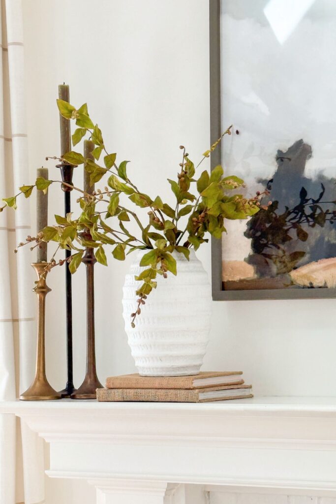 White textured vase with green branches styled on a mantel with brass candlesticks, stacked books, and framed art