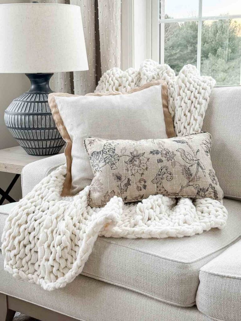 Chunky knit throw layered with neutral pillows on a light upholstered sofa in a cozy winter living room.
