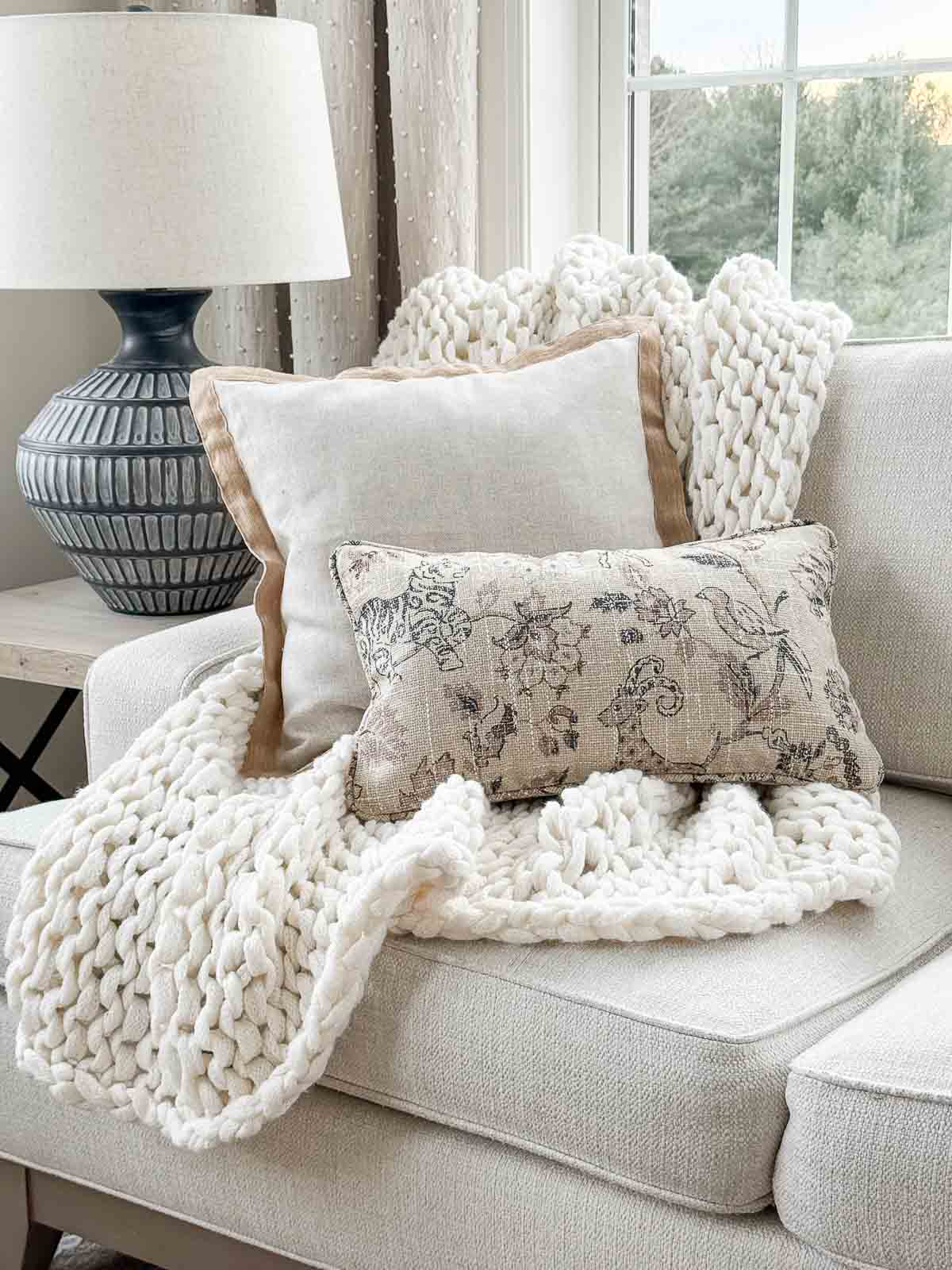 Chunky knit throw layered with neutral pillows on a light upholstered sofa in a cozy winter living room.