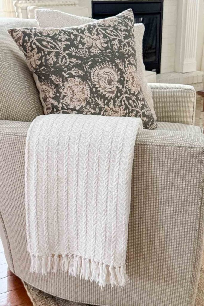 Neutral upholstered chair styled with a cream knit throw and a soft patterned pillow in a cozy winter living room.