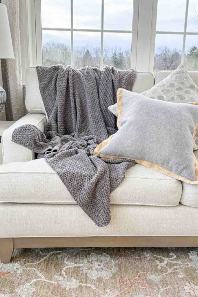 Neutral throw pillows layered with a textured gray throw on a light upholstered sofa in a cozy winter living room.