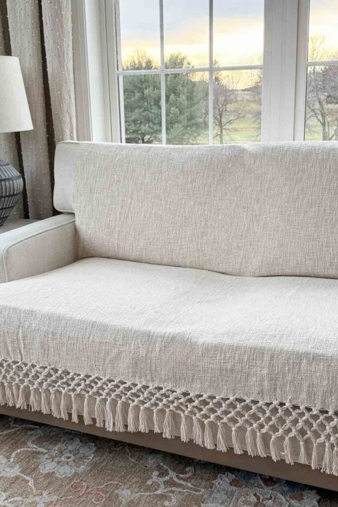Neutral sofa layered with a lightweight fringed throw in a bright winter living room with large windows.