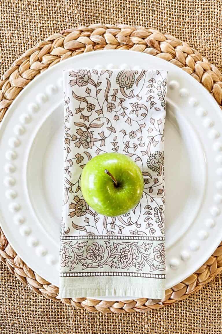 Green apple on a floral napkin atop white beaded plates for weekly menu plan post