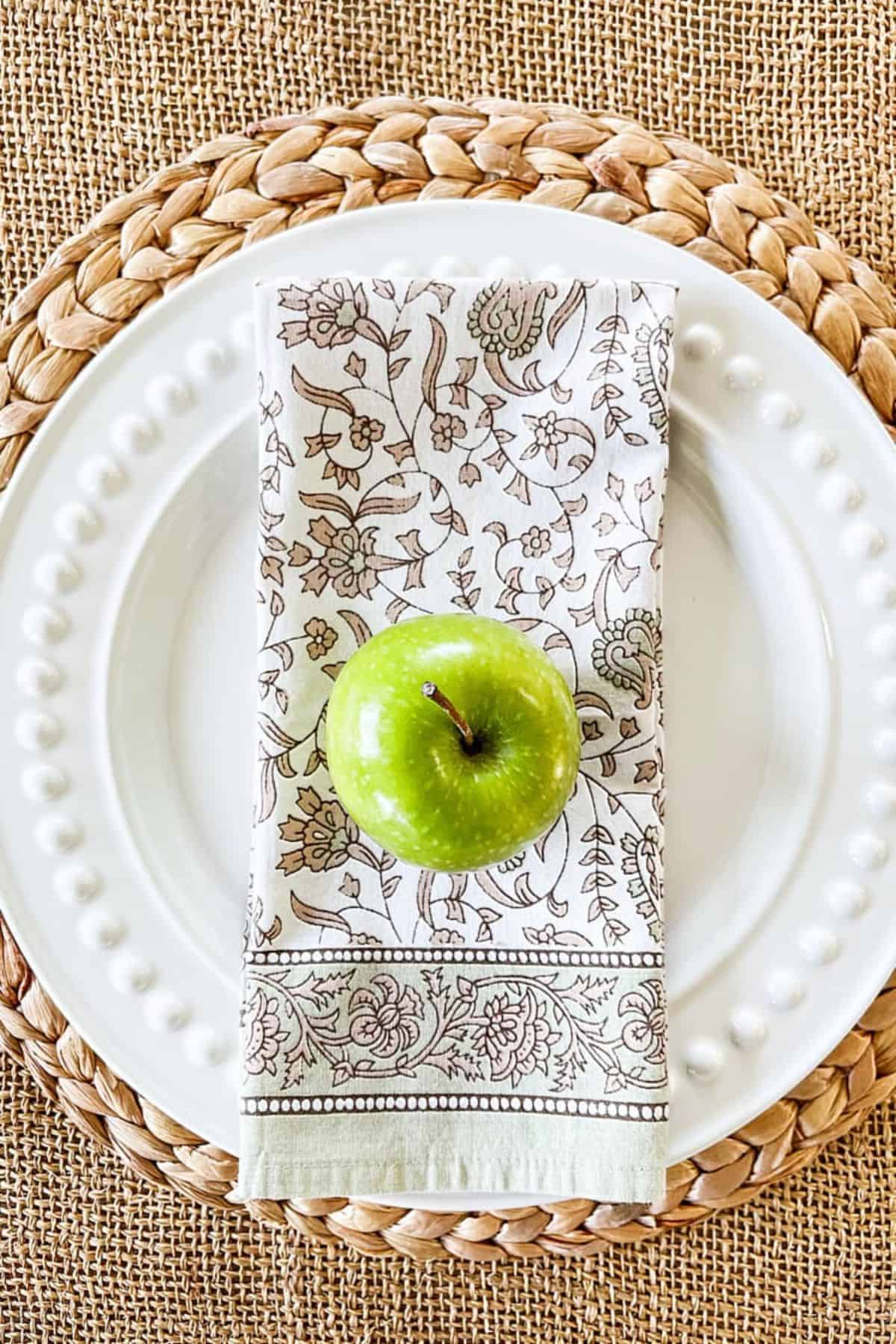 Green apple on a floral napkin atop white beaded plates for weekly menu plan post