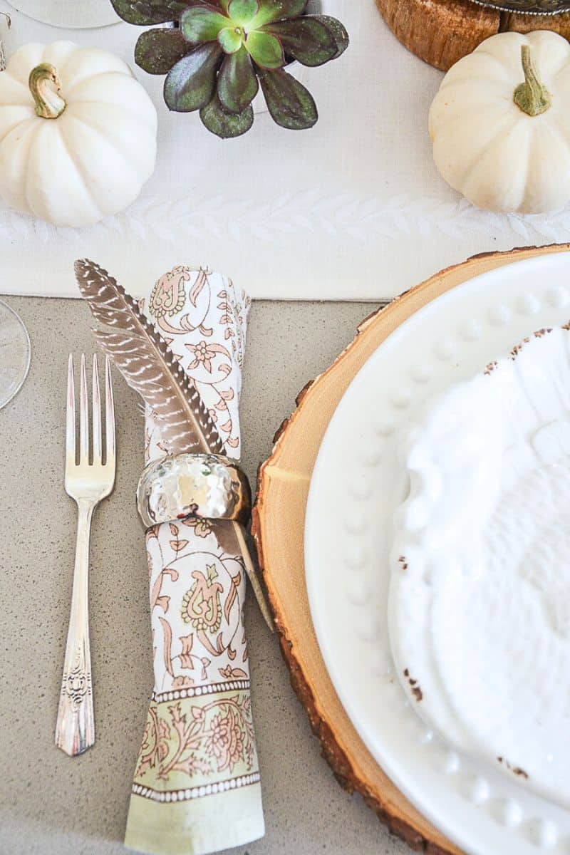a pretty fall inspired napkins rolled up in a silver napkin ring with a small pheasant feather tuck into it.
