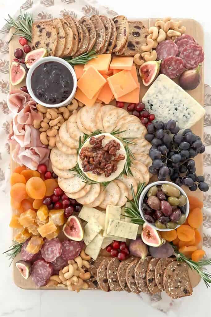 Holiday charcuterie board arranged with cheeses, crackers, nuts, dried fruit, olives, grapes, and fresh rosemary on a wooden board.