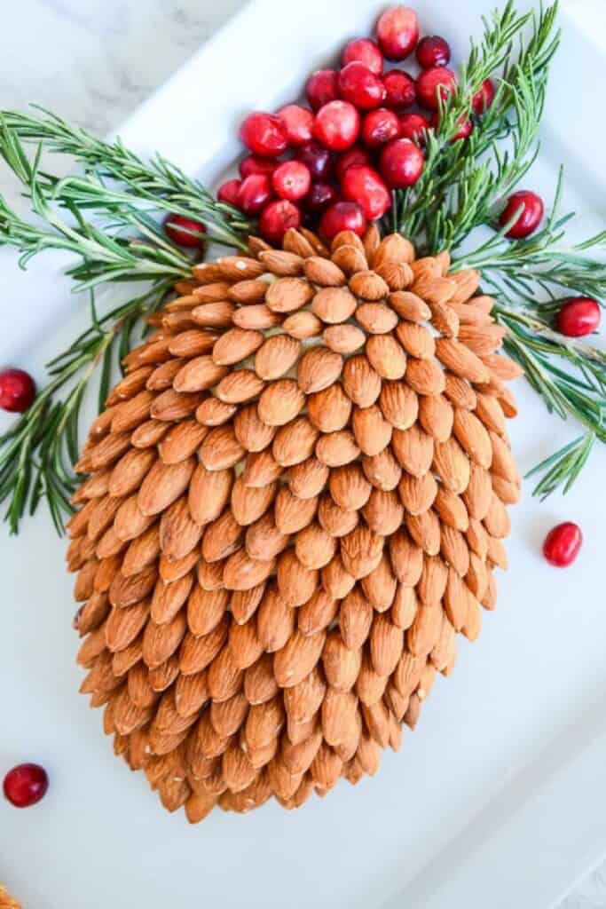 Almond covered pinecone cheese ball garnished with rosemary and cranberries on a white platter.