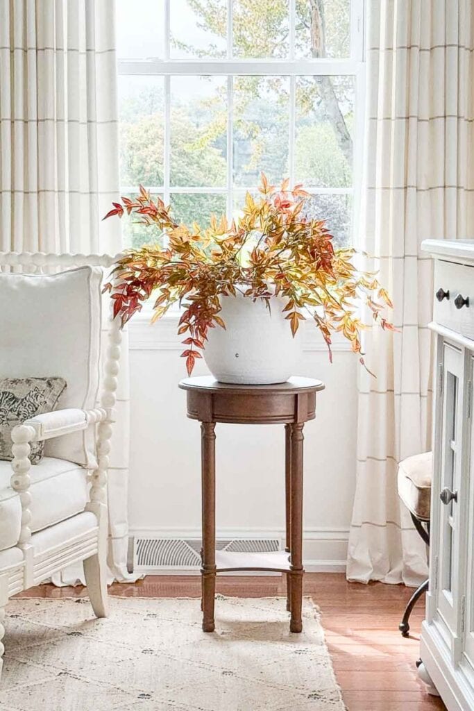 Bright living room corner with a white spindle chair, neutral pillow, and a wood pedestal table holding a white vase of autumn branches.