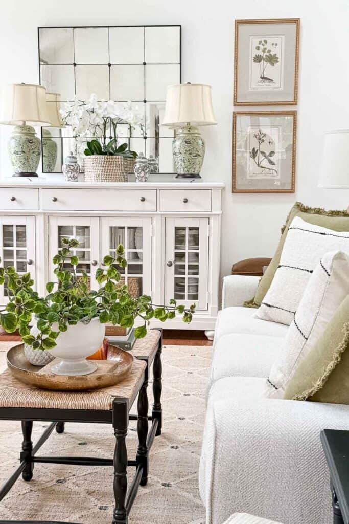 White buffet styled with green ceramic lamps, large square mirror, botanical prints, and a woven planter with orchids for layered decorating details.