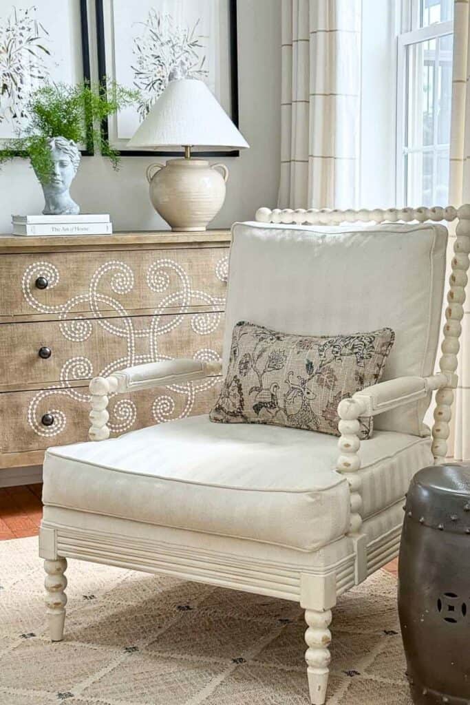 White spindle chair with patterned pillow beside a decorative chest styled with books, a lamp, and a bust planter with greenery.