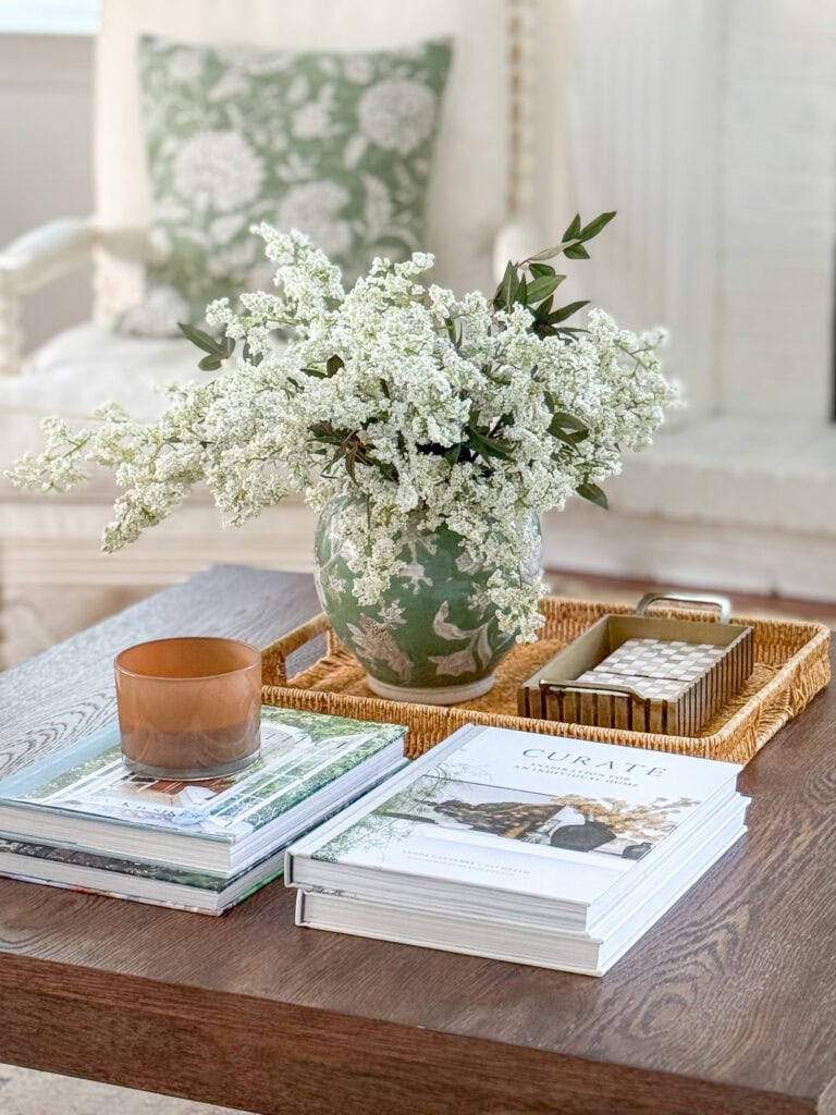 green ginger jar with white flowers styled on a wicker tray coffee table vignette with decorating books and a green floral pillow in the background