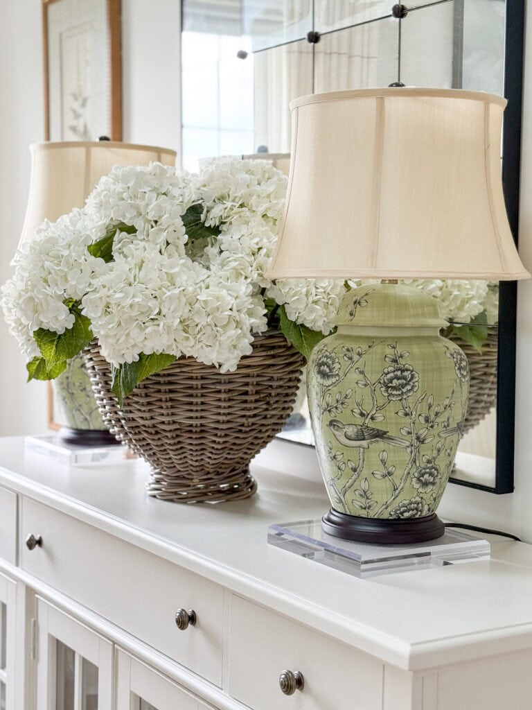 faux white hydrangeas in a wicker basket next to a green chinoiserie lamp on a white sideboard