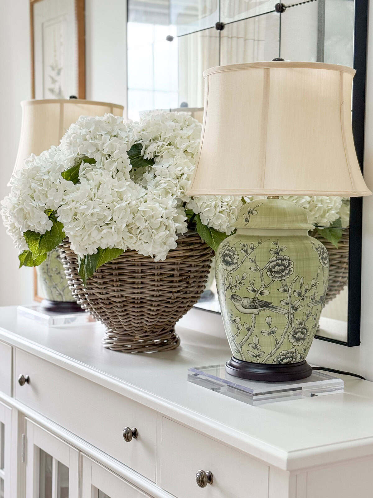 faux white hydrangeas in a wicker basket next to a green chinoiserie lamp on a white sideboard