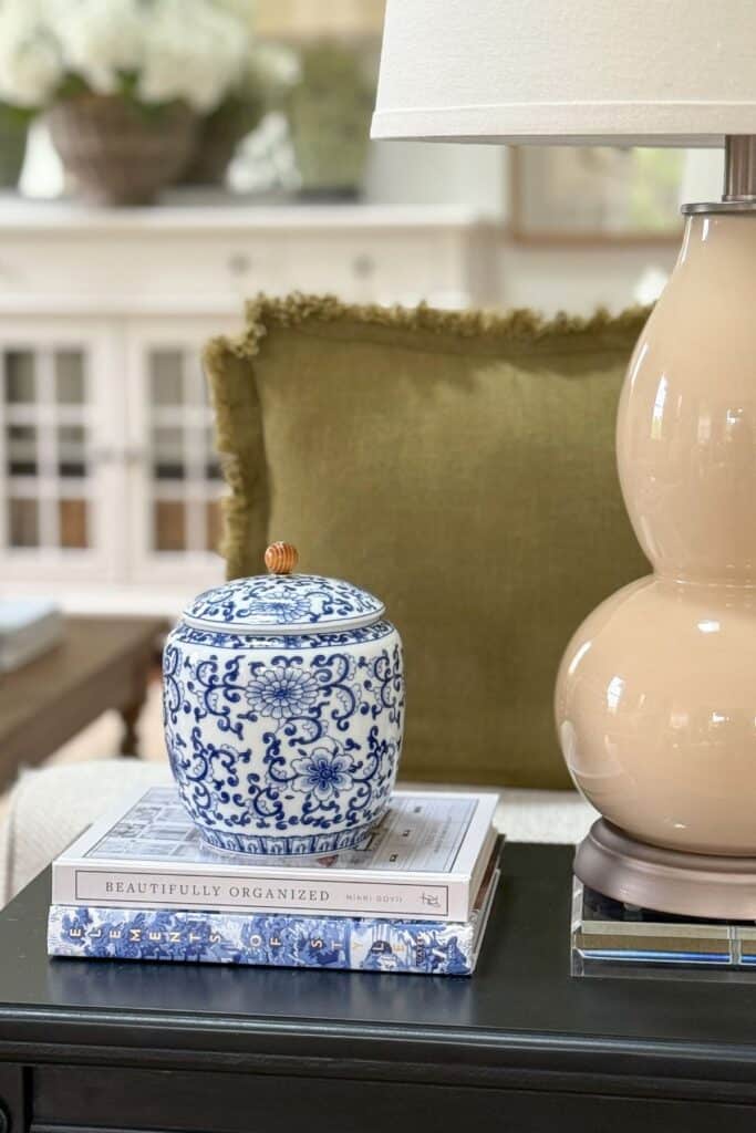 blue and white ginger jar stacked on books on a dark side table with a green pillow and neutral lamp in a neutral living room