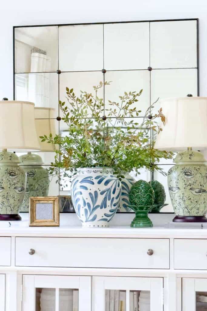 green chinoiserie lamps and blue painted vase with green branches styled on a white neutral sideboard with an antique mirror