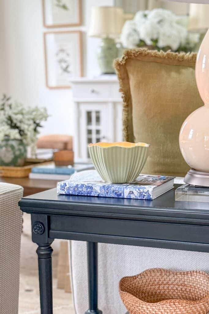green scalloped bowl and blue book styled on a dark sofa table with a cream lamp and green velvet chair in a neutral living room