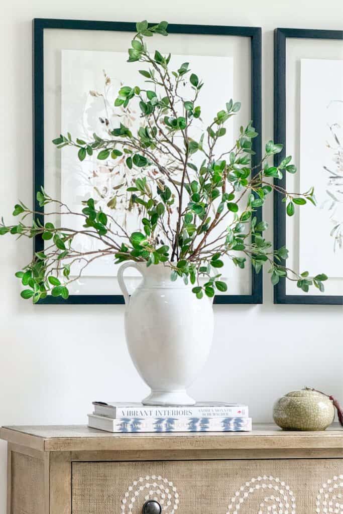 green branches in a white ceramic vase styled on a neutral console table with framed art and decorating books