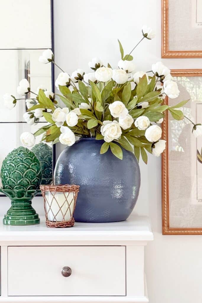 Rule of three vignette with blue vase of white ranunculus, green ceramic artichoke, and woven candle on a white dresser