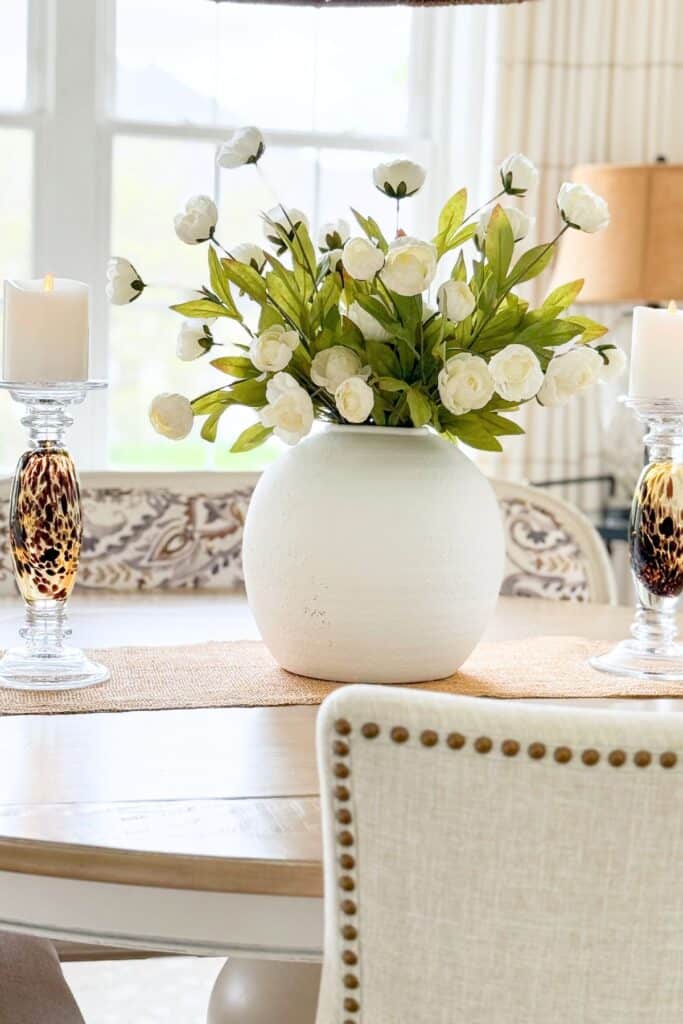 white faux flowers with green stems in a white round vase on a neutral dining room table with candles