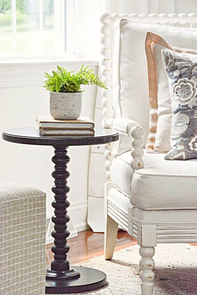 small drink table styled with books and plant next to upholstered chair with pillows