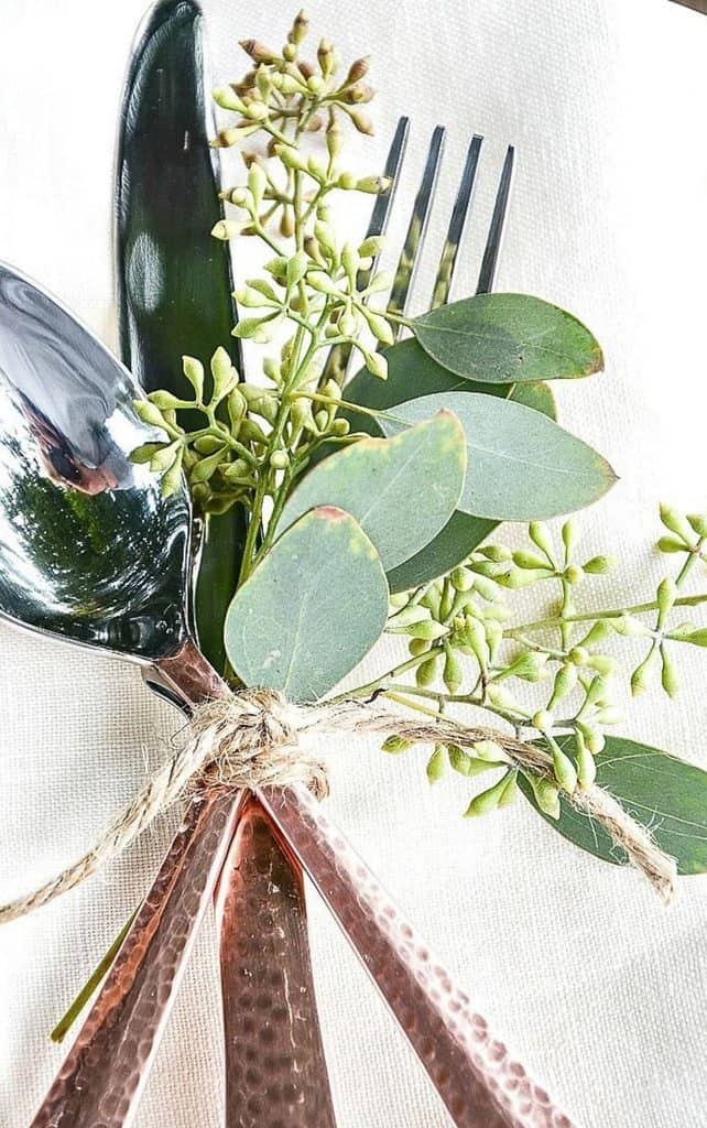 Hammered copper flatware tied with twine and a sprig of seeded eucalyptus on a white linen napkin for a fall place setting