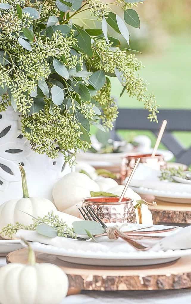 Outdoor fall table with seeded eucalyptus in a white pierced vase, white pumpkins, hammered copper mugs, and white plates on log slice chargers with copper flatware tied in twine