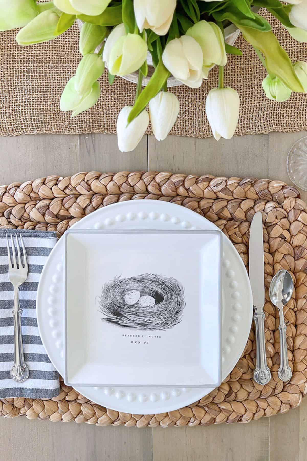 spring table setting with white tulips, woven placemat, white dishes, and silver flatware