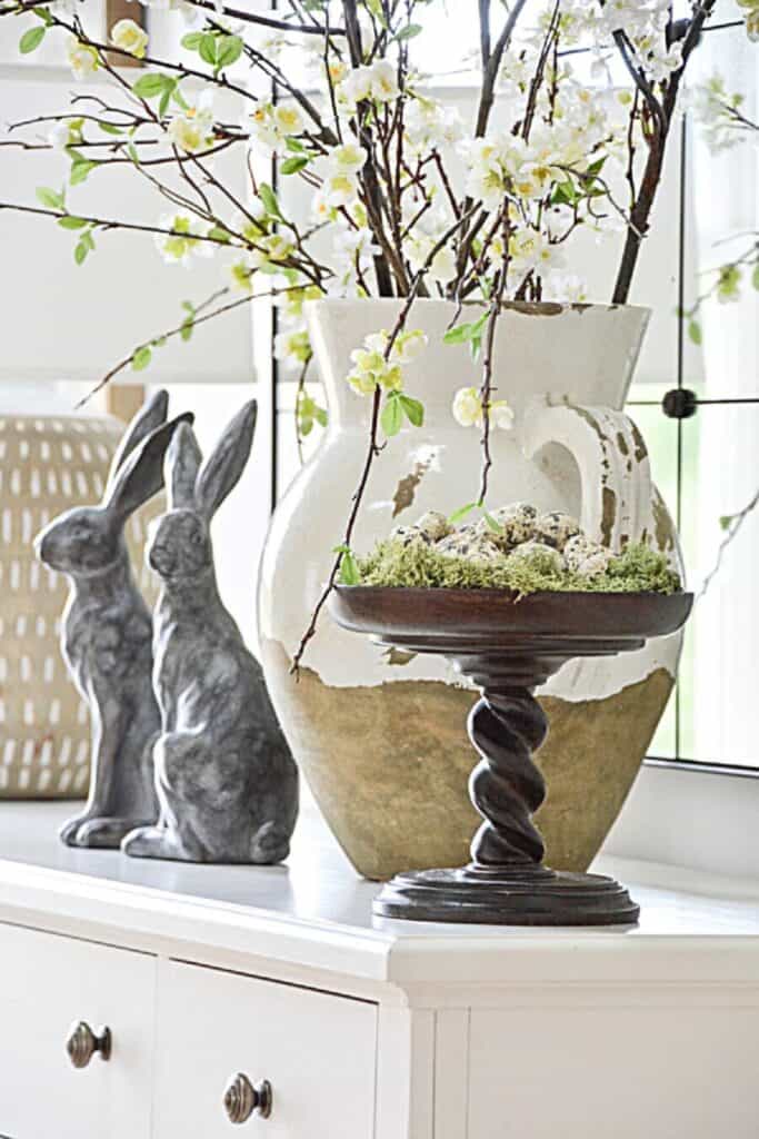 after Easter spring vignette with blooming branches in a large vase, moss, eggs in a bowl, and decorative rabbits on a cabinet