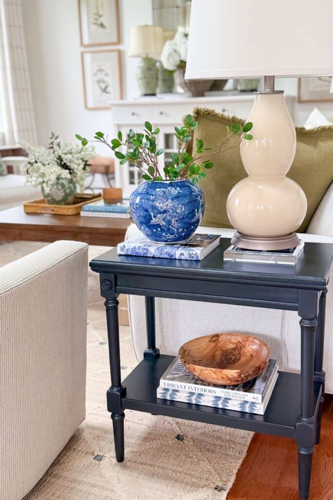 Beige living room with a black side table styled with a blue and white vase, cream lamp, and carved wood bowl