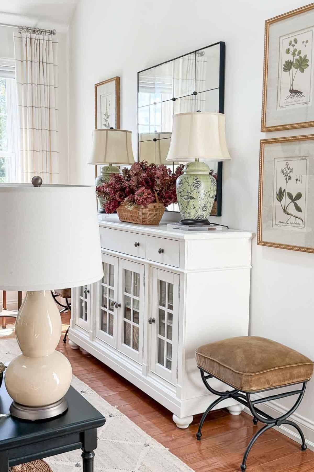 Warm neutral living room with a white cabinet, camel stool, green ginger jar lamps, and dried burgundy hydrangeas