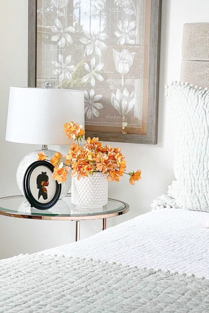 Realistic faux orange flowers arranged in a white vase on a bedroom nightstand