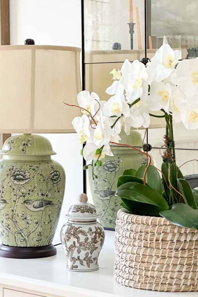Realistic faux white orchids styled in a woven basket on a white dresser with decorative jars